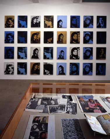 A grid of canvases in shades of blue and gold hanging on a wall. The repeated image is of Jackie Kennedy, wife of former President John F. Kennedy. In the foreground is a vitrine displaying various magazine and newspaper clippings surround the JFK assassination.