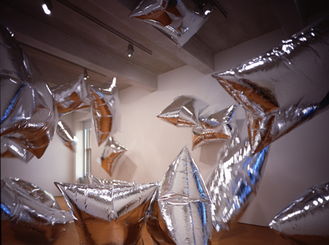 Retangular silver foil balloons float in an empty white-walled room with a brown floor.