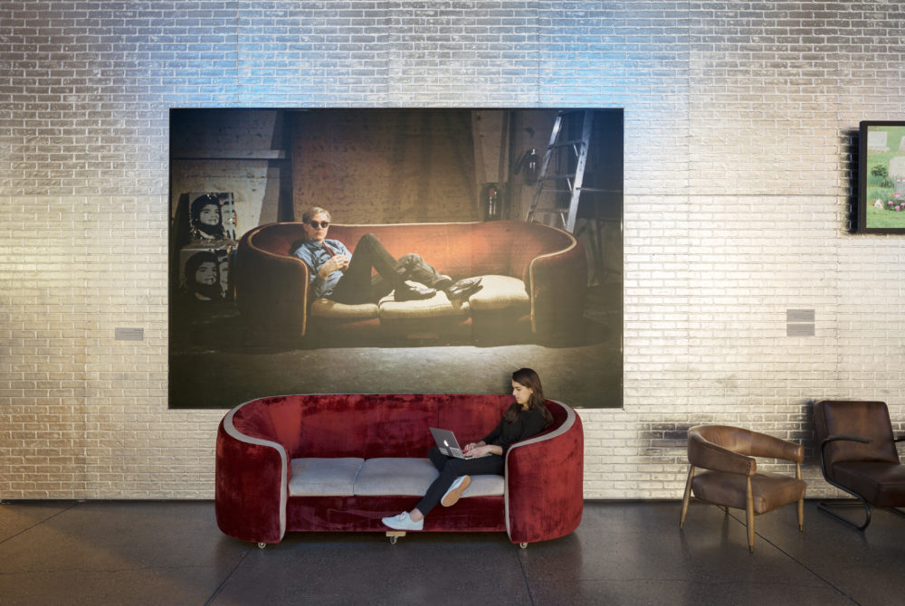 A young woman reclines on a red velvet couch in the Andy Warhol Museum Lobby, working on an Apple laptop, under a portrait of Andy Warhol reclining on a red velvet couch.