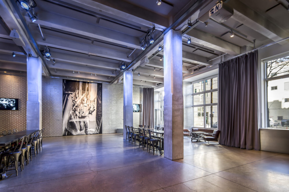 A photograph of the Andy Warhol Museum entrance space. The far wall is silver brick and a large picture of Andy Warhol holding up a Marilyn print. Large windows on the right side of the image look out to the street. There are large square pillars in the space, as well as long tables surrounded by chairs.
