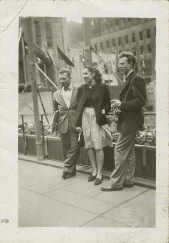 A young Andy Warhol stands outdoors with two other people alongside a railing.