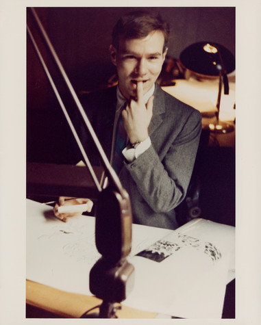 A photograph of And Warhol working at a desk covered in a large sheet of white paper. He is wearing a grey suit and has his left index finger in his mouth as he gazes toward the camera.