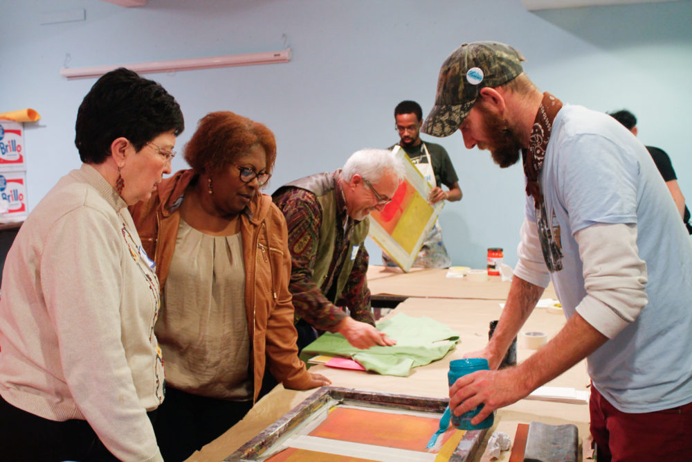 An artist educator spreads blue ink on a screen as he prepares to help two older women create their own screen prints.