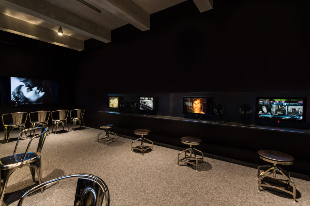 The film and video gallery in the Andy Warhol Museum, where visitors can sit and explore the Warhol's film and video collection at their leisure using touch screens. Silver chairs line the perimeter of the room in front of monitors and a projected screen test on the far wall.