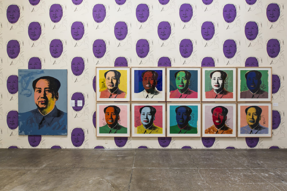 Eleven Mao portraits in different colors hanging on a wall wallpapered with purple Mao portraits.