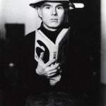 Andy Warhol holds an open book and looks directly at the viewer in a promotional photoshoot.