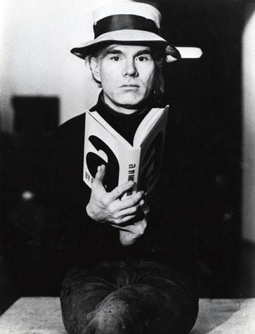 Andy Warhol holds an open book and looks directly at the viewer in a promotional photoshoot.