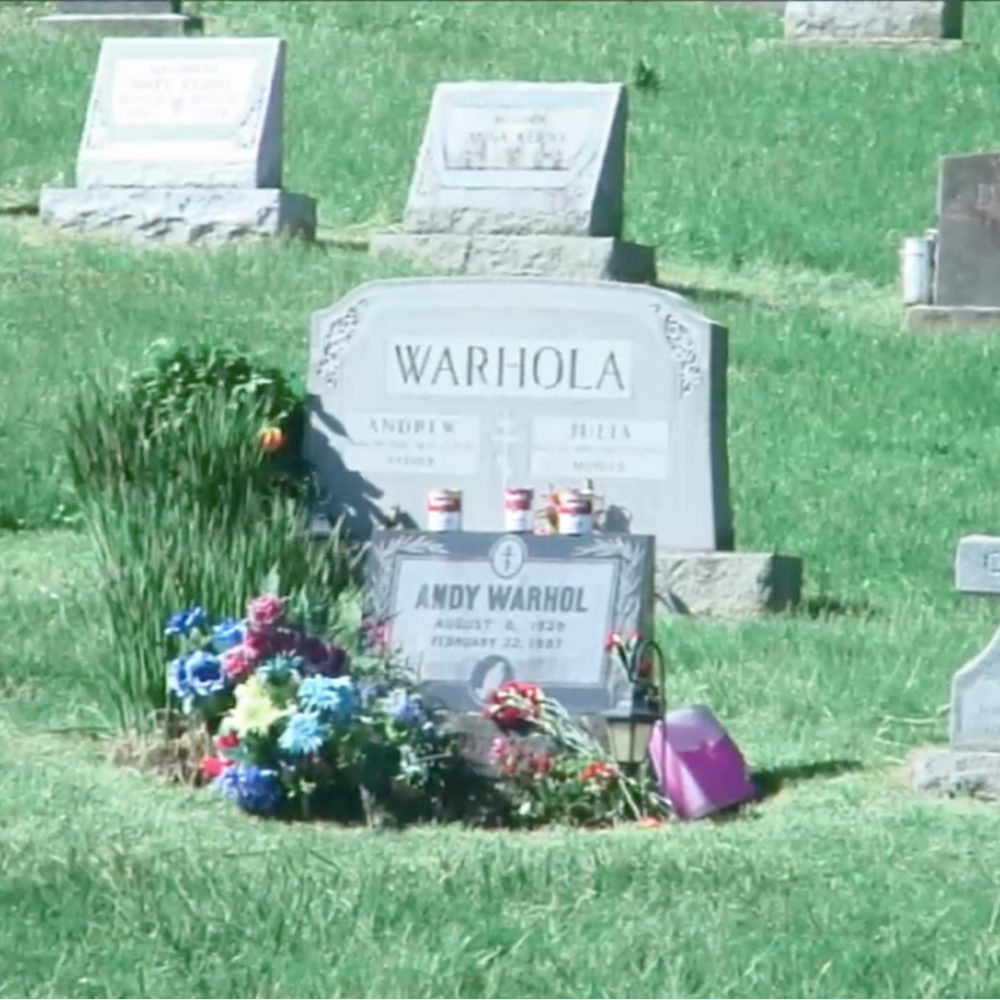 A photograph of Andy Warhol's grave in the springtime. There are flowers around the headstone and Campbell's soup cans placed on top of it