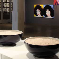 In this installation shot from the exhibition Andy Warhol | Ai Weiwei, two oversized bowls filled with pearls sit on a white pedestal, with portraits by Andy Warhol visible in the background.