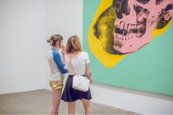 Two teens look at an oversized painting of a skull rendered in pastel pink, seafoam green, and bright yellow