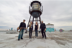 Four members of Hypercube standing on a councrete building roof in front of a water tower behind them, also on the roof. From left to right, a man in a hat holds a guitar, a woman holds a saxaphone, a women stands arms to her sides, a man stands holding percussion mallets.