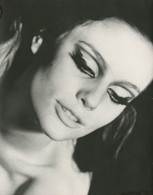 A black and white headshot of a female model wearing heavy eye makeup. The picture has been captured at an angle, and the woman’s face tilts down toward the right corner of the image.