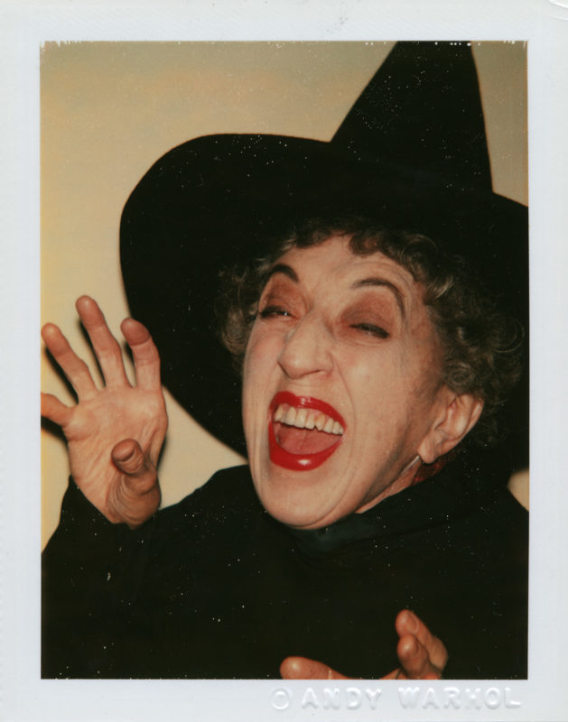 A woman wearing white face powder, bright red lipstick, and a black witch’s hat poses with her hands raised, mouth opened as though she is cackling.
