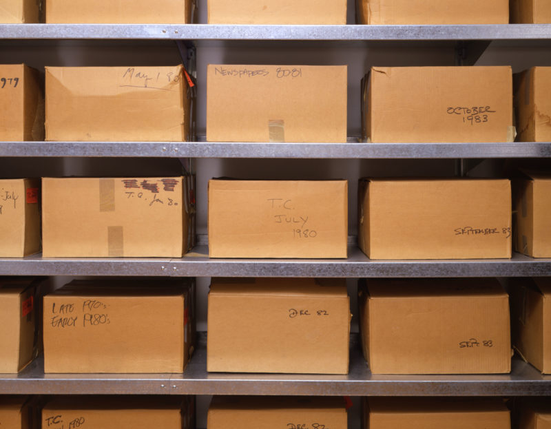 This image depicts Andy Warhol's time capsules as they are displayed in the Warhol archives. Neat rows of brown cardboard boxes, all the same size, are lined up on metal shelves. On some of the boxes, tape is visible, or there is writing in black sharpie noting the date that the time capsule was sealed.