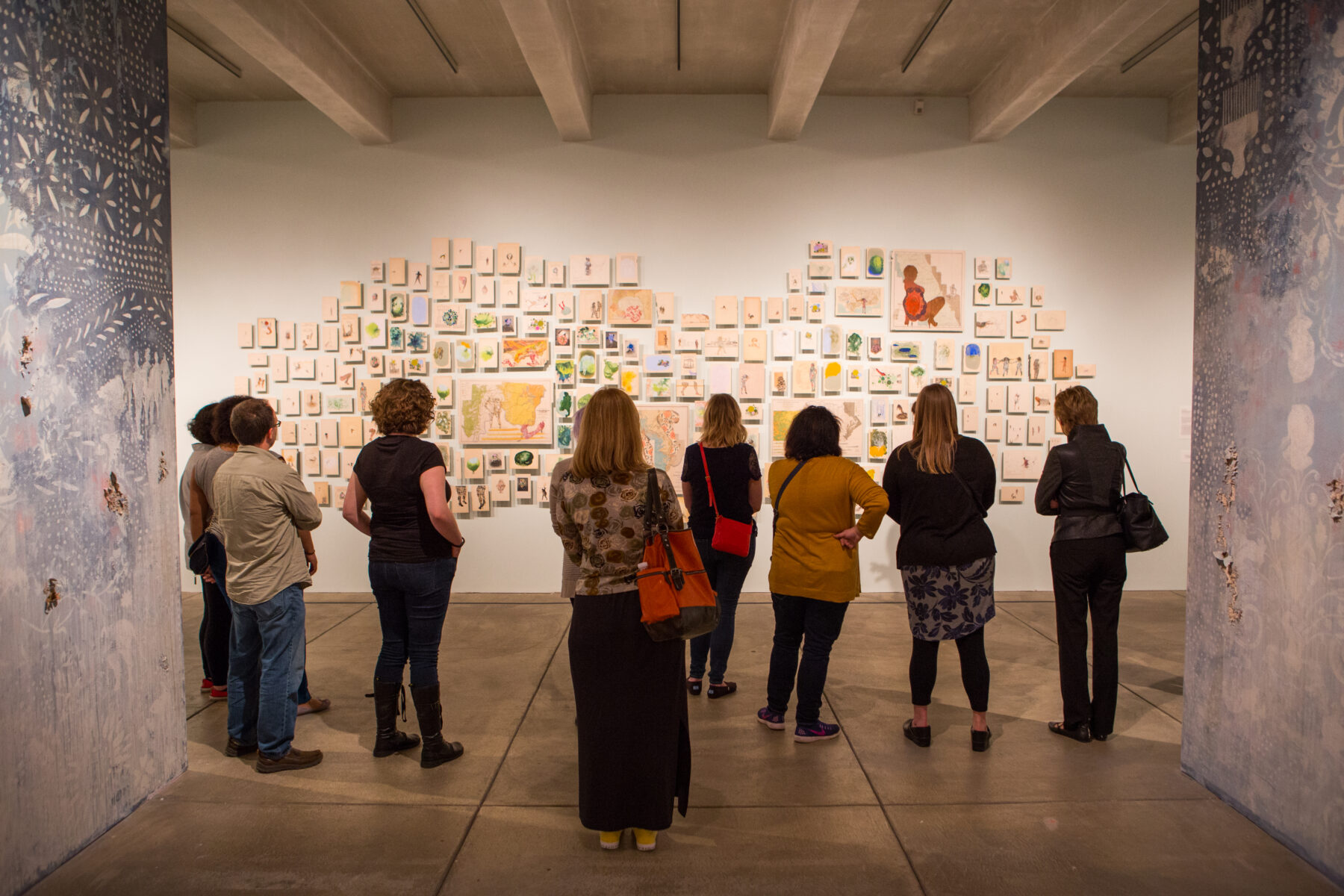 A group of visitors face away from the camera as they examine a display in the Firelei Baez bloodlines exhibition. Small paintings of various sizes are tacked to the wall in a large cluster in front of them.