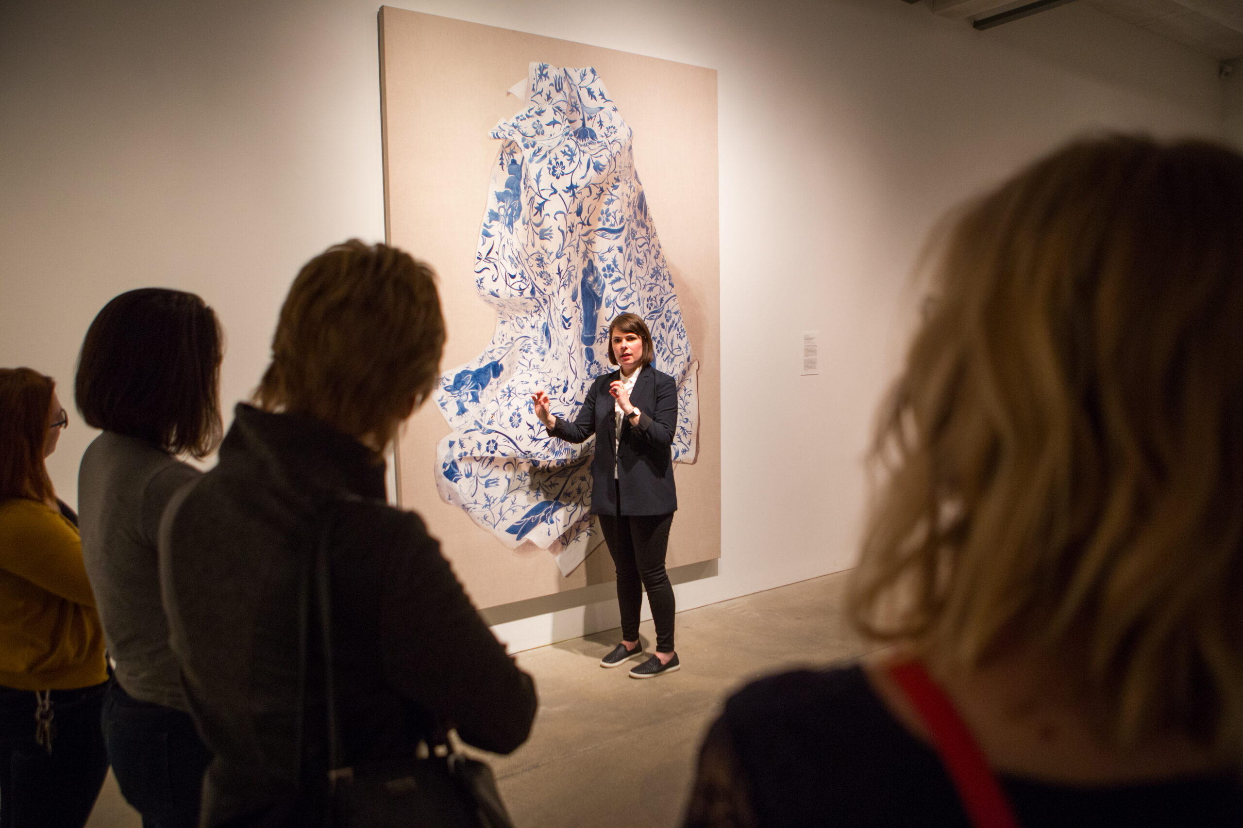 A woman with short brown hair gestures towards a large painting of fabric decorated with delicate blue designs at a Sip and Sketch event.