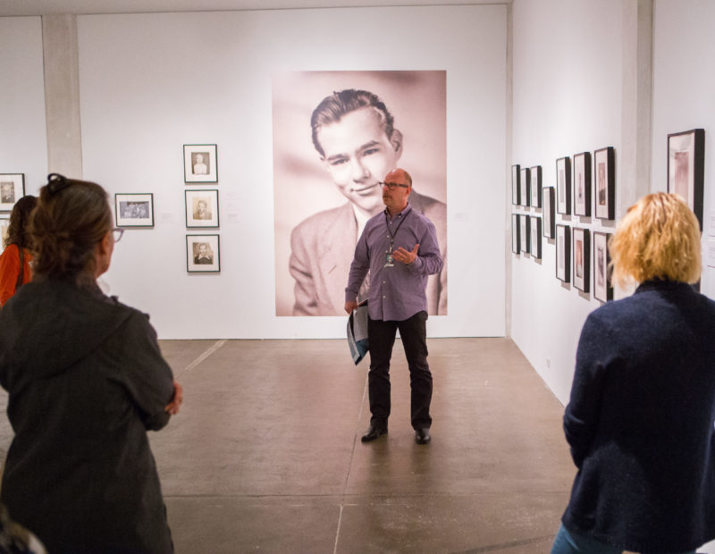 A man in a purple button-up shirt leads a group of educators through a gallery during teacher open house.