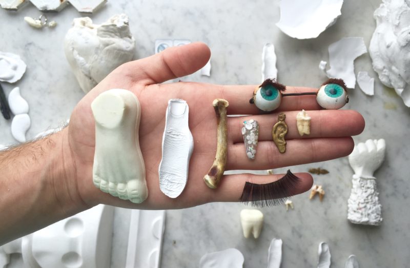 In this installation shot of Adam Milner's art, a hand holds various small objects representing body parts, including doll eyes, a small bone, and a plaster imprint of a finger.