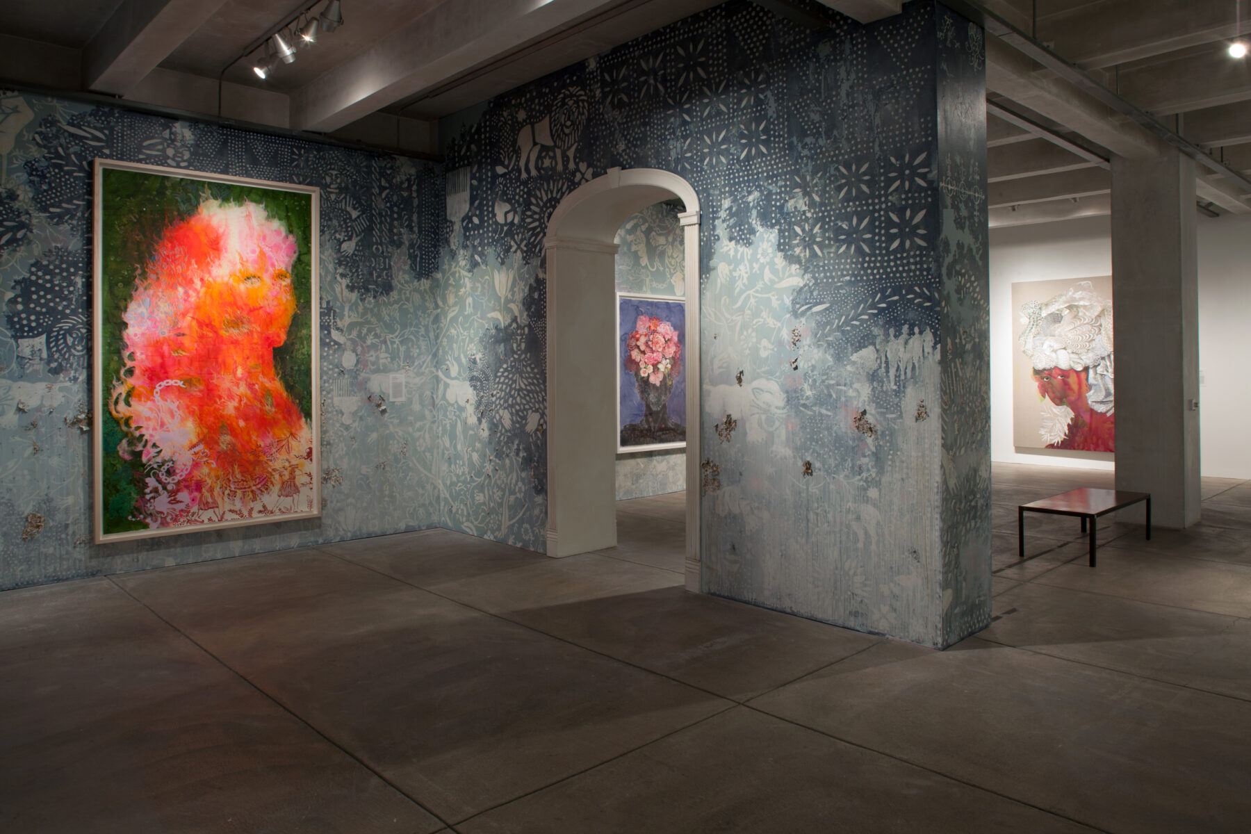 In this photograph of the Bloodlines exhibit, the center of the image is taken up by a wall decorated with ornate blue designs and an arch in the center. Several paintings of women in profile are visible.