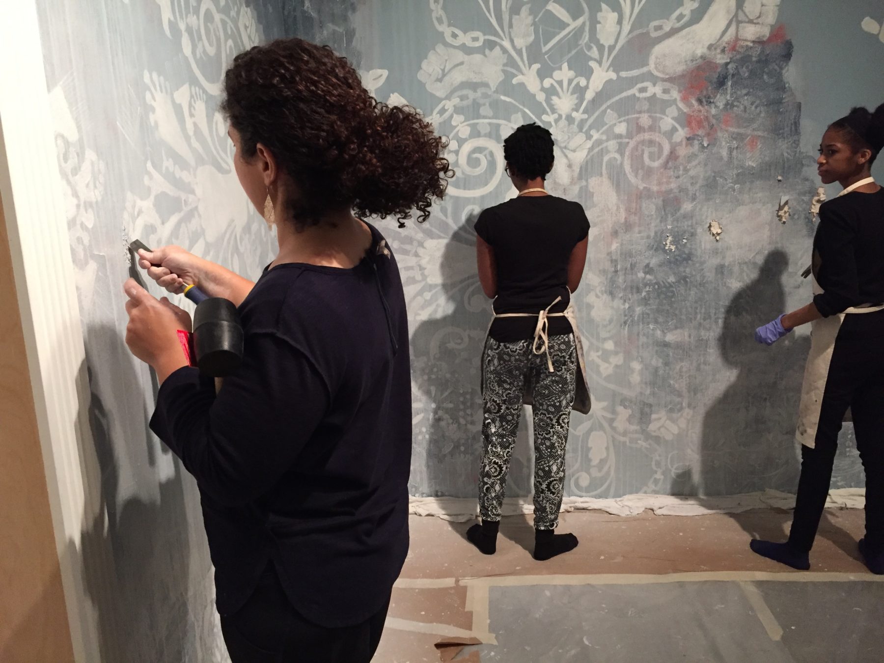 A group of volunteers help artist Firelei Baez scrape paint off a wall in her exhibition gallery.