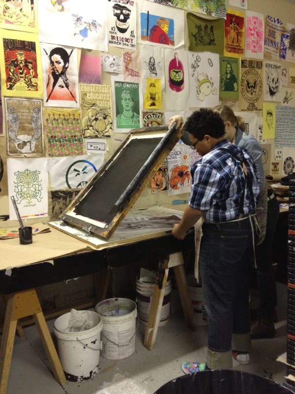 Two artists work on creating a print at a table in front of a wall filled with prints made by other artists.
