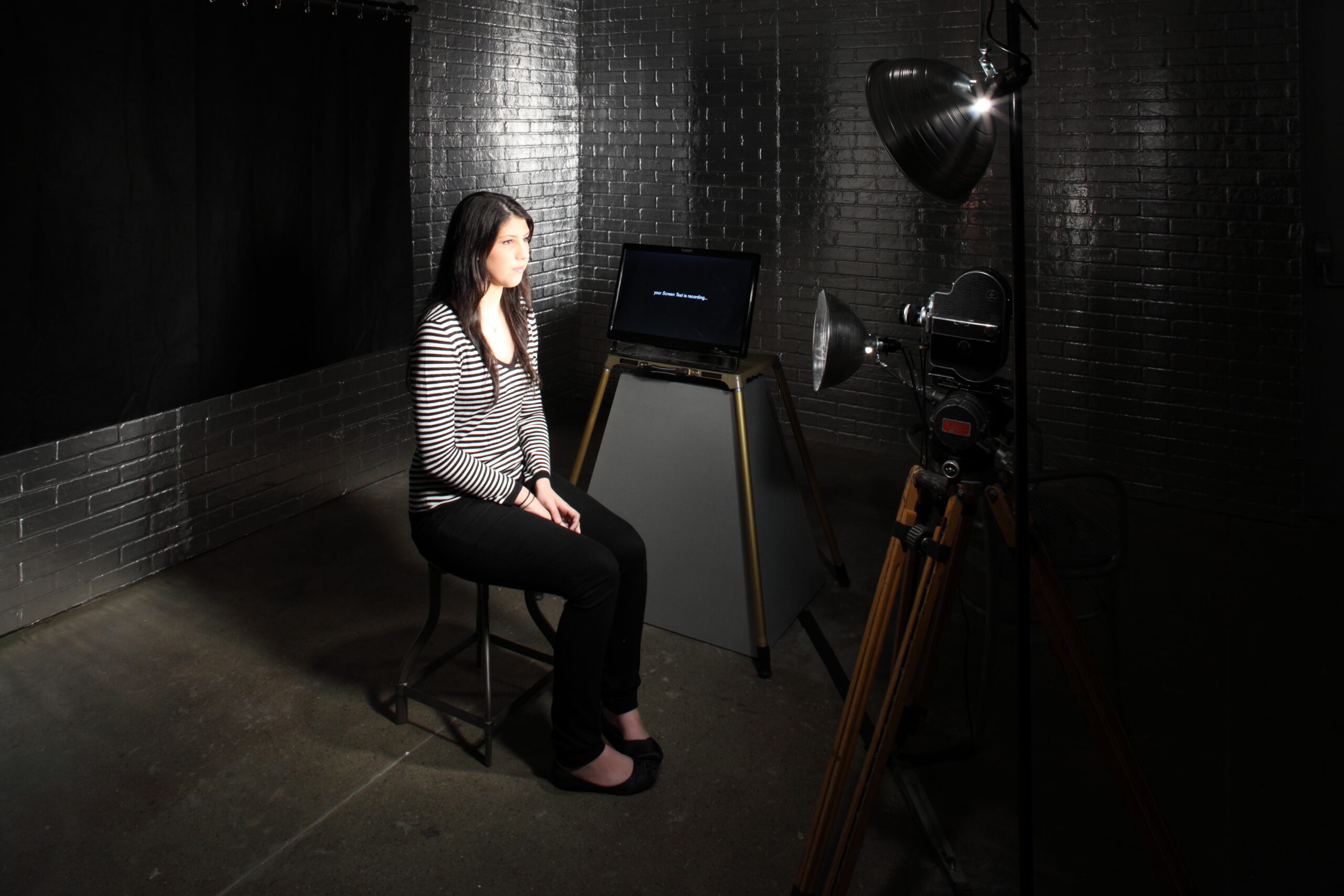 In a room with silver brick walls, a woman sits in front of an old-fashioned camera.