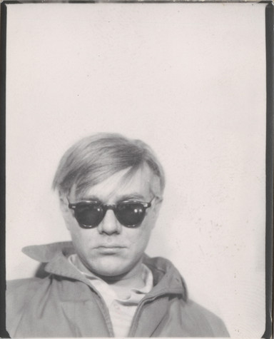 In this black and white photograph, Andy Warhol stands against a blank background wearing a hooded jacket and dark sunglasses, an indifferent expression on his face.