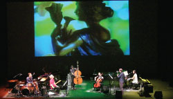 An orchestra is seated on stage. Behind them is a video projection showing a classic sculpture against a blue and green lit background.