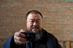 Artist Ai Weiwei stands against a brick backdrop and holds a point-and-shoot camera in front of him