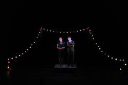 Two performers stand side-by-side on a dimly lit stage, surrounded by blackness with only a string of multi-colored holiday lights framing them.