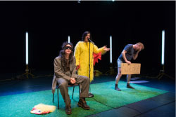 Three performers are on a sparsely decorated stage. The man on the left of the image is seated, wearing a brown suit, black blindfold over his eyes, and long brown hair. The woman in the center is standing behind a microphone with her left hand raised, and she is wearing a yellow chicken suit (The head of the suit is on the floor next to the seated man.). The man on the right is wearing socks, boxers, and a t-shirt, holding in his right hand a cardboard sign that says algebra, and his head is in his left hand in a sign of exasperation.