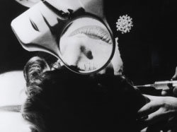 Black-and-white film still showing a woman's head resting on a table with her face looking up. A magnifying glass held in front of the face magnifiys her nose and lips. In the lower right of the scene is a hand holding a syringe to the woman's temple.