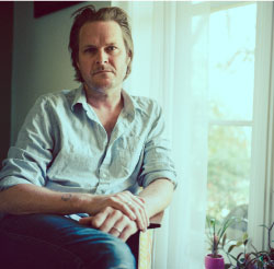Caucasian man sits legs crossed and right hand on top of his left wrist, resting on his legs. His button-up shirt is a bit wrinkled with sleeves rolled up, and he is wearing jeans. He looks directly into the camera with a straight face. He is seated in front of a window.