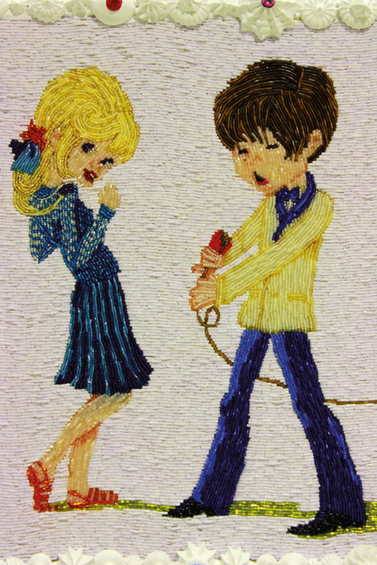A detail of an artwork depicting a girl with blond hair, wearing a blue dress and red shoes with a blue bow in her hair and a boy with short, brown hair with a yellow jacket on and blue pants holding a microphone. The boy appears to be singing in the microphone. The artwork is comprised of glaze, hand embroidered beads, paint, and plastic pearls.