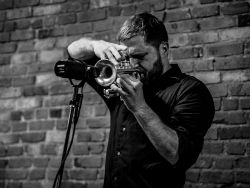 Black-and-white image photograph of a bearded man playing a trumpet into a microphone. A brick wall is behind him.