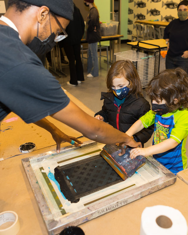 Photograph of two children creating a screenprint with the help of another person in The Factory studio of The Andy Warhol Museum.