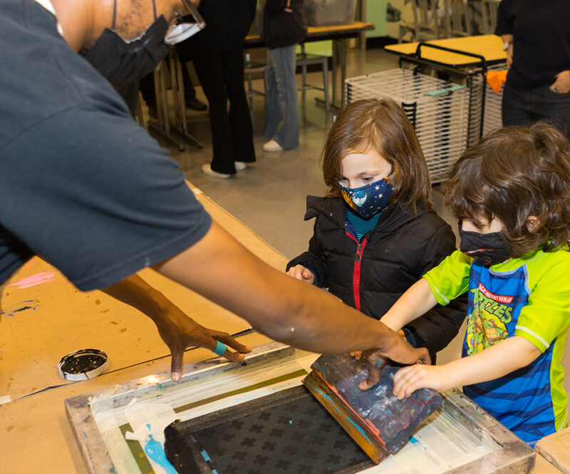 Photograph of two children creating a screenprint with the help of another person in The Factory studio of The Andy Warhol Museum.