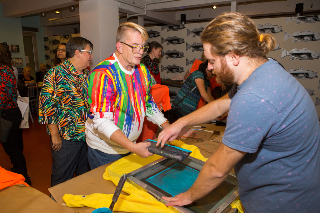 An artist educator helps a teacher wearing glasses and a white sweatshirt with colorful streaks on it screen print a design onto a yellow shirt using turquoise ink.