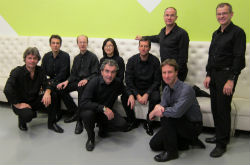 Nine people dressed in black seated and standing around a white couch.
