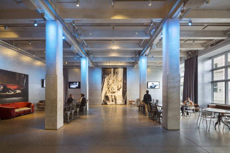 A photograph of the Andy Warhol Museum entrance space. A red velvet couch sits against the left side of the image with a photograph of Andy Warhol on a red velvet couch on the wall behind it. Large windows and round metal tables with chairs around them are to the right. In the center of the image there are four cement pillars highlighted with blue lighting, which have long rectangular tables between them. A photograph of Andy Warhol holding a portrait of Marilyn Monroe is against the far wall.