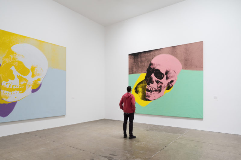 A man in a red hoodie stands with his hands on his hips as he examines a massive screen print of a skull in The Andy Warhol Museum.
