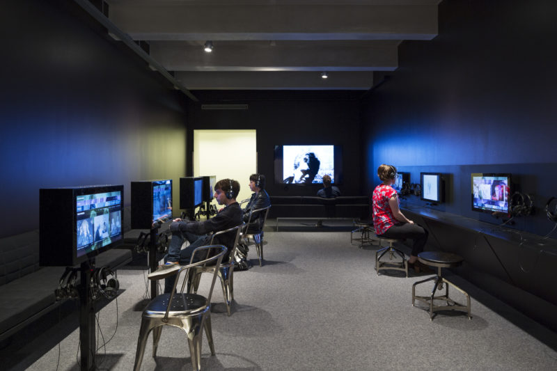 Visitors wearing headphones interact with devices housing Andy Warhol’s film and video works lining the walls of a room with black walls. A visitor watches a video that is projected on the back wall of the room.