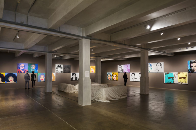 A photograph of a gallery with cement floors and grey walls that are covered in large pairs of screen-printed portraits. Four pillars in the center of the room surround a few pieces of furniture draped in white fabric.