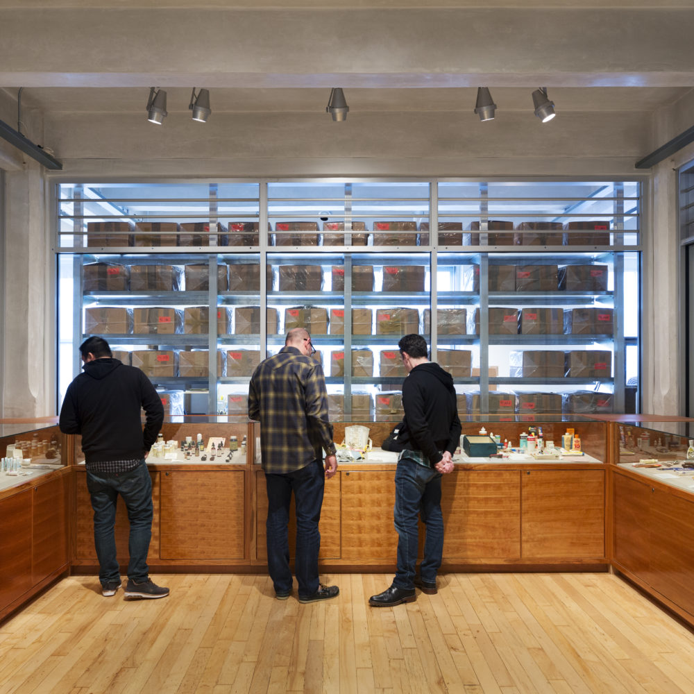 Three men examine artifacts in the Andy Warhol Museum Archives. Three walls of the room have glass cases, and through the glass wall rows and rows of the cardboard boxes that are Andy Warhol’s time capsules are visible.