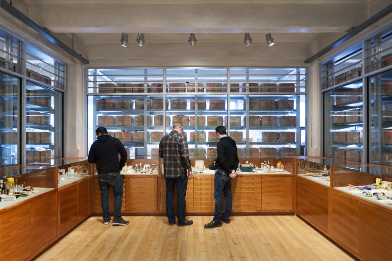 Three men examine artifacts in the Andy Warhol Museum Archives. Three walls of the room have glass cases, and through the glass wall rows and rows of the cardboard boxes that are Andy Warhol’s time capsules are visible.