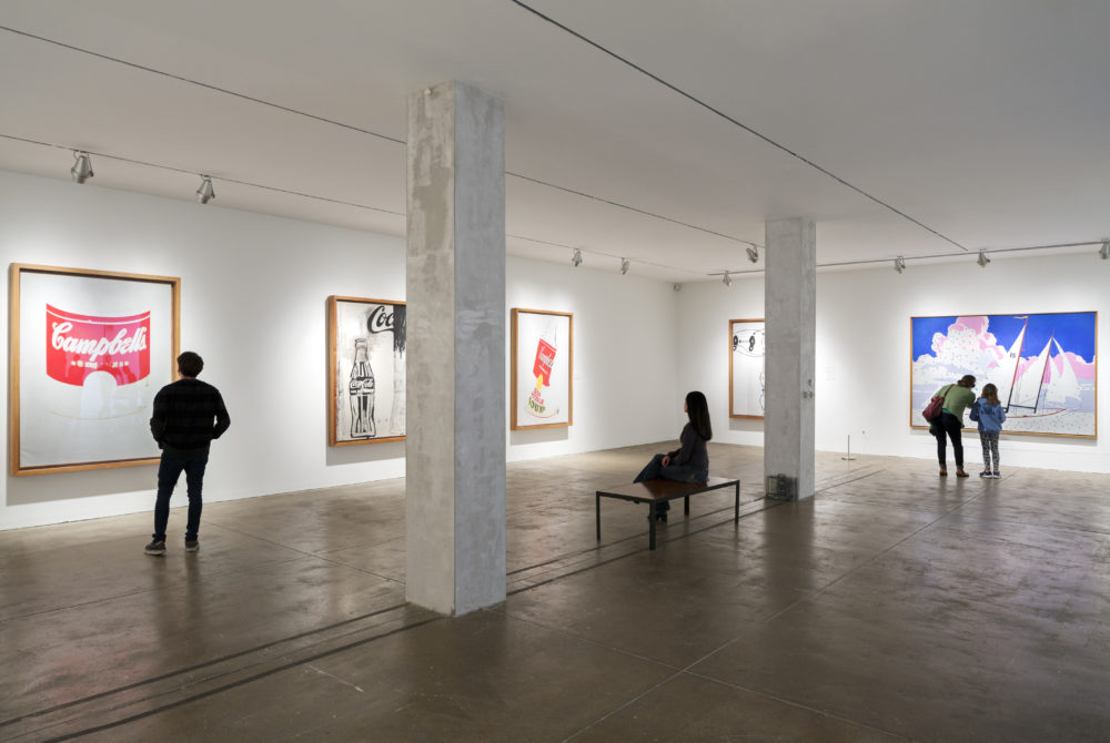 One of The Andy Warhol Museum’s galleries. The walls and ceiling are white, the floor is cement, and two large pillars are in the middle of the room. A male visitor examines a hand-painted Campbell’s soup can. A woman sits on a bench and looks at a picture featuring a Coca-Cola bottle. A woman and a child examine a painting of a sailboat.