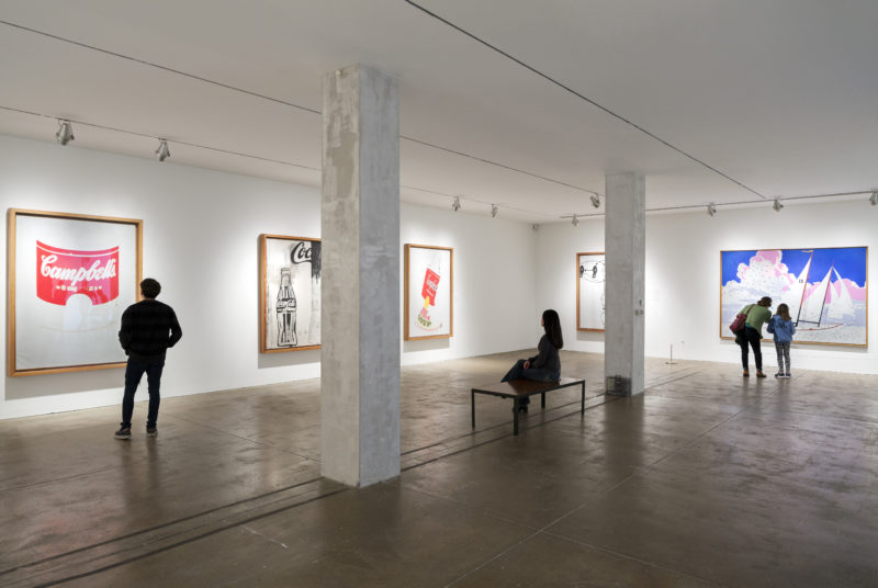 One of The Andy Warhol Museum’s galleries. The walls and ceiling are white, the floor is cement, and two large pillars are in the middle of the room. A male visitor examines a hand-painted Campbell’s soup can. A woman sits on a bench and looks at a picture featuring a Coca-Cola bottle. A woman and a child examine a painting of a sailboat.