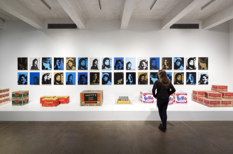 A woman with brown hair dressed entirely in black faces a display of Andy Warhol’s works. Two rows of small, screen-printed portraits are hung on a white wall. Some of Warhol’s sculptural work-- including reproductions of Campbell’s boxes, Brillo boxes, and a tray of silver Coca-Cola bottles-- sit on a low platform in front of the portraits.