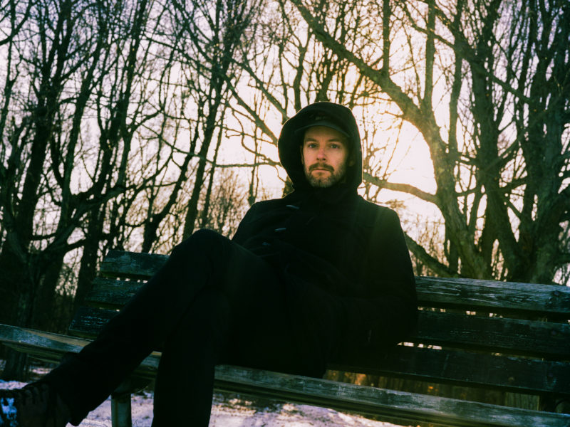 Photograph of a man seated on a wood park bench. He wears all black and has a hood pulled over his head. Trees in the background have no leaves, and the sun is setting directly behind the seated man.