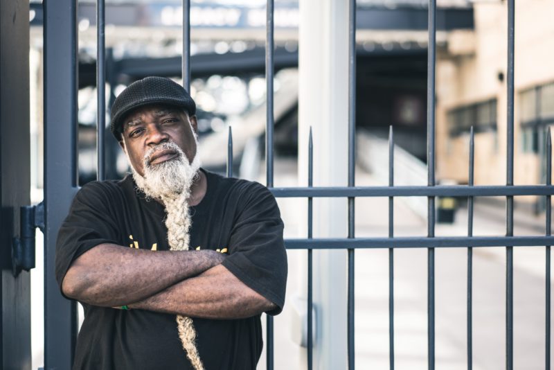 African American man leaning against a fence, looking directly at the viewer, arms folder across his chest. He has a long white beard in a braid and is wearing a black hat and black t-shirt.
